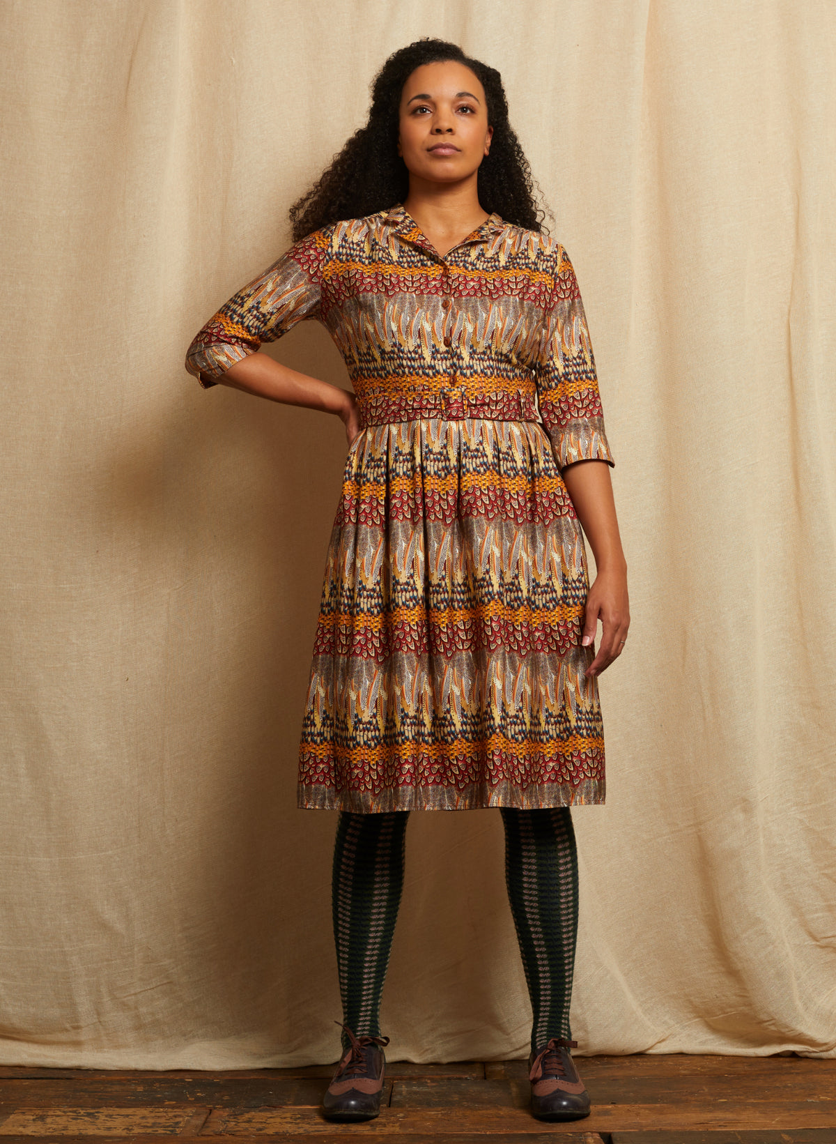 A woman stands in front of a beige fabric backdrop wearing a patterned collared dress with three-quarter sleeves, a fitted waist, and a flared skirt. The dress has an intricate multicolor geometric and tribal-inspired feather print in beige, orange, red, blue, and brown.