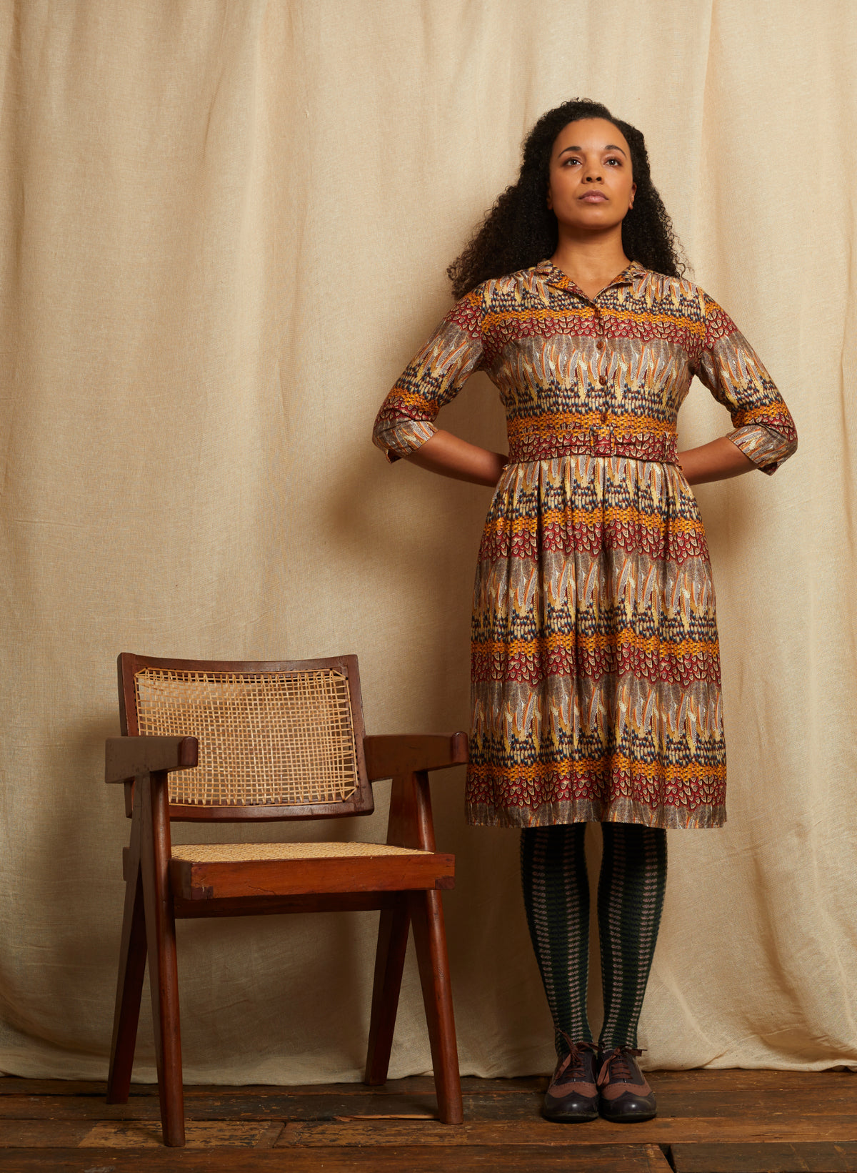 A woman stands in front of a beige fabric backdrop wearing a long-sleeve collared dress with a multicolor geometric and tribal-style featherpattern in red, orange, yellow, and brown. She is wearing dark green and black striped tights and dark shoes.