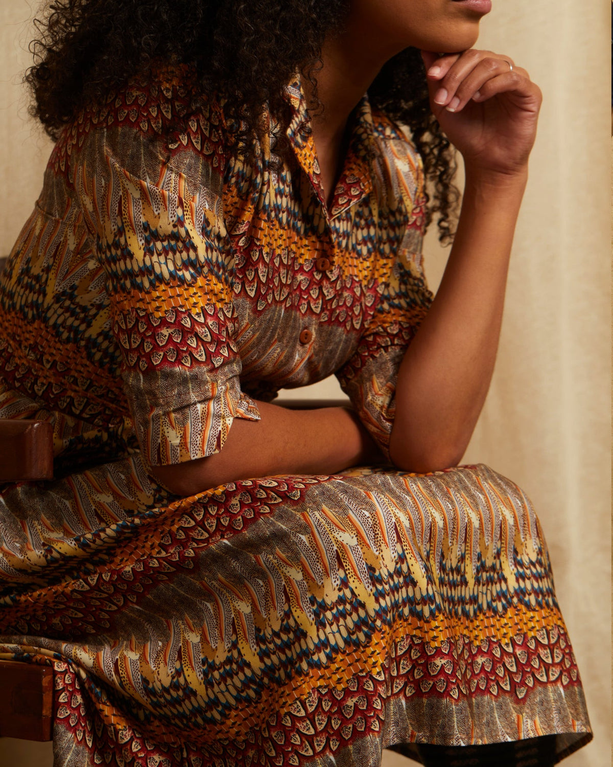 A woman wearing a patterned kaftan feather dress with a geometric and floral design in brown, gold, red, and blue, sitting against a neutral beige background.