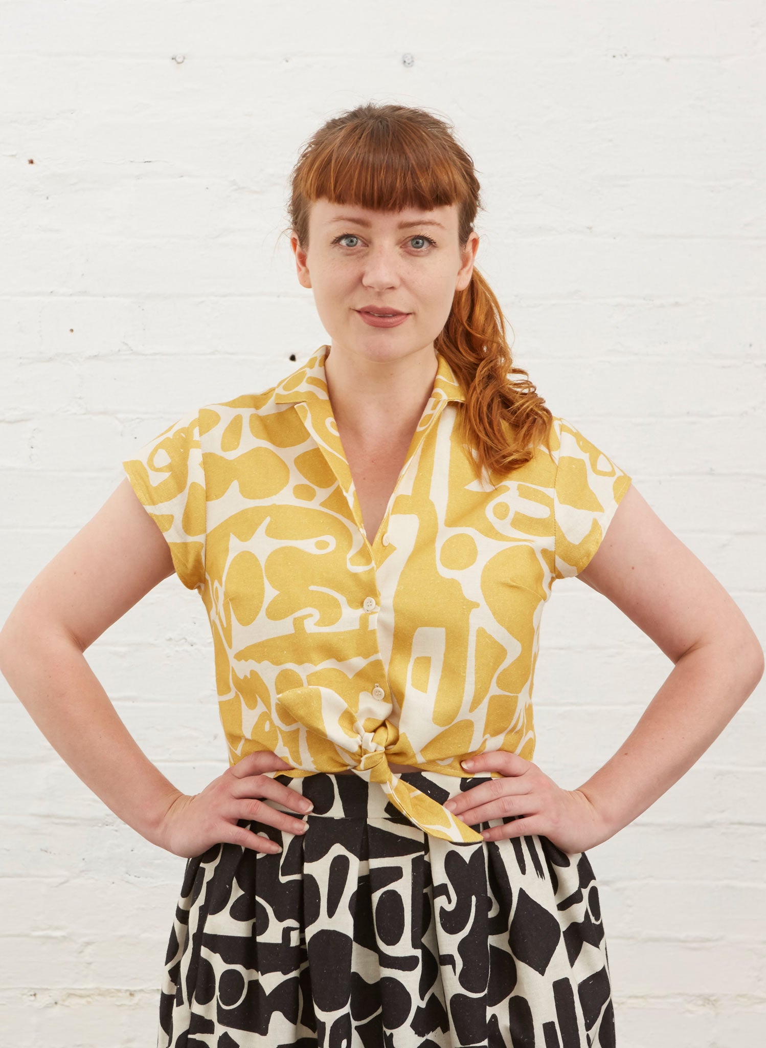 Photo of Alison wearing a cropped tie waist button up blouse with yellow and white abstract shape fabric. Also wearing black and white Ada skirt in same pattern.