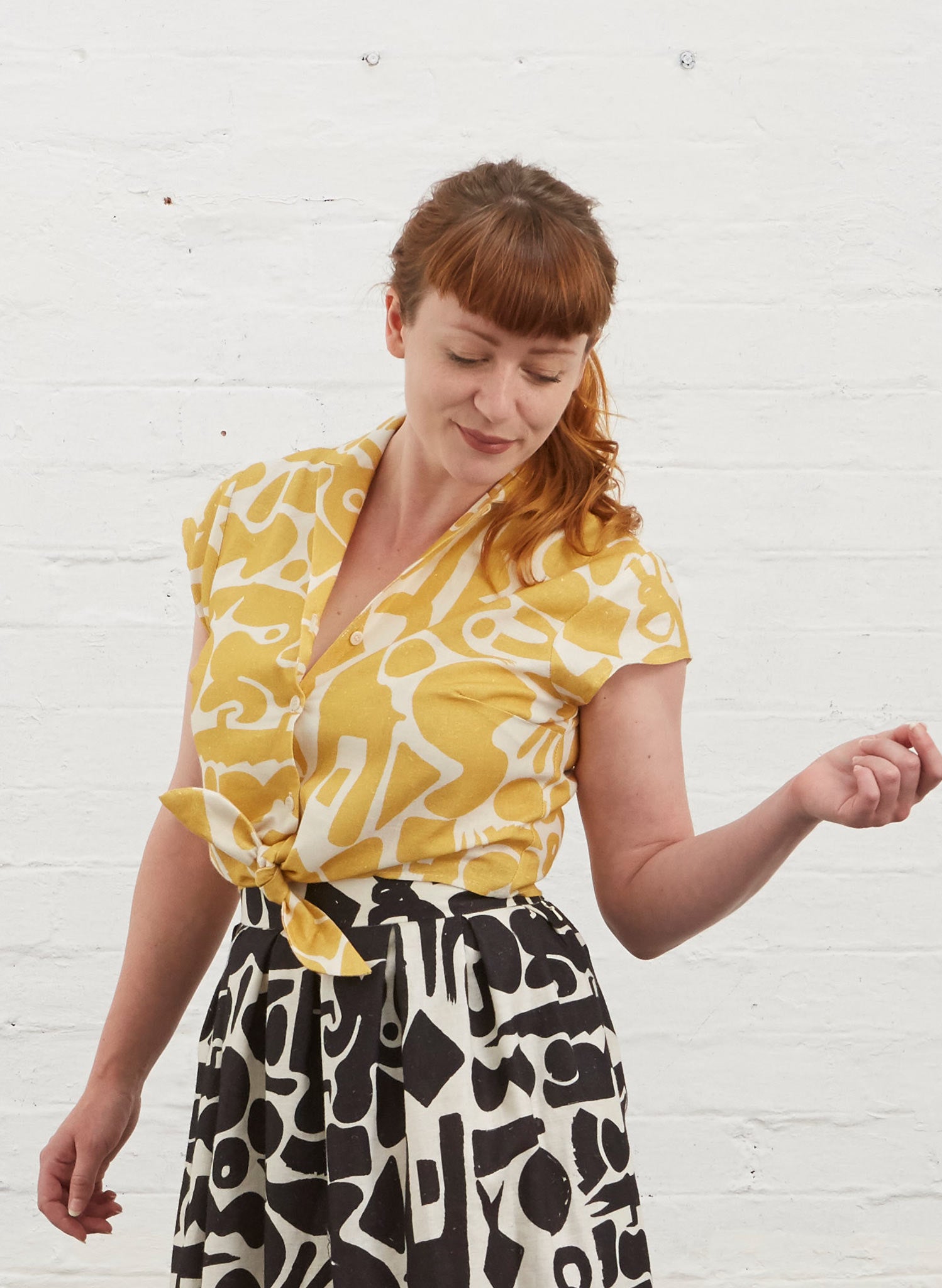 Photo of Alison ( fringed auburn hair )wearing a cropped tie waist button up blouse with yellow and white abstract shape fabric. Also wearing black and white Ada skirt in same pattern.